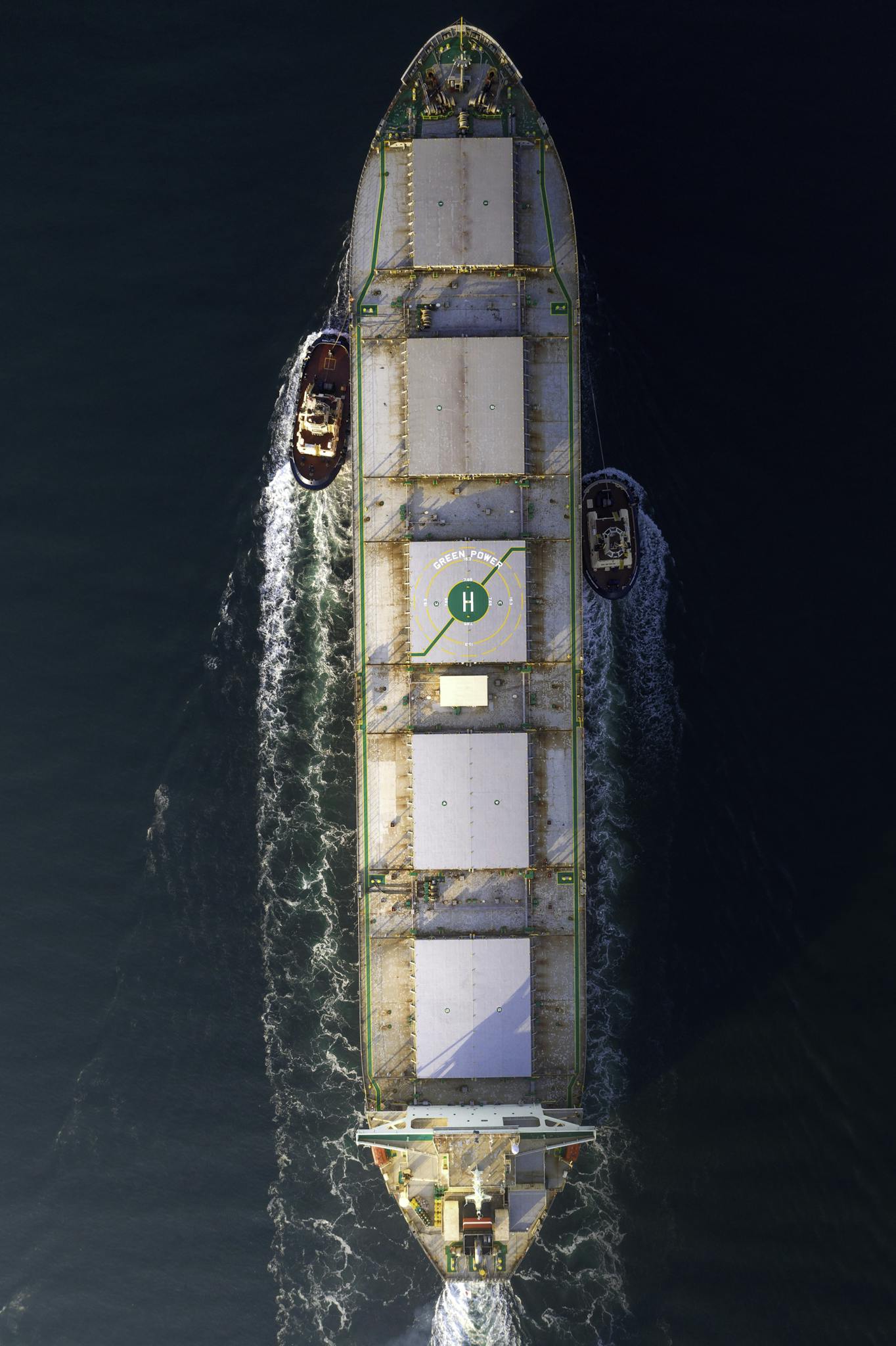 An aerial photo of a large cargo ship with tugboats assisting in open sea, showing maritime transportation.
