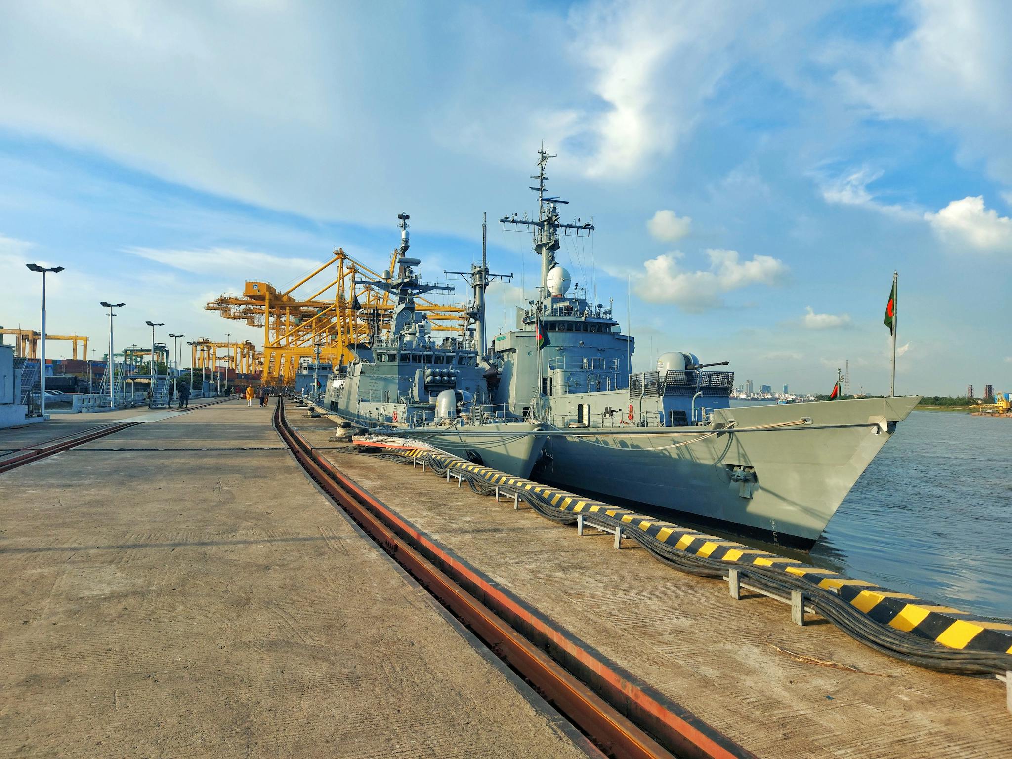 Navy vessels docked at a bustling city port with cranes and clear blue skies.
